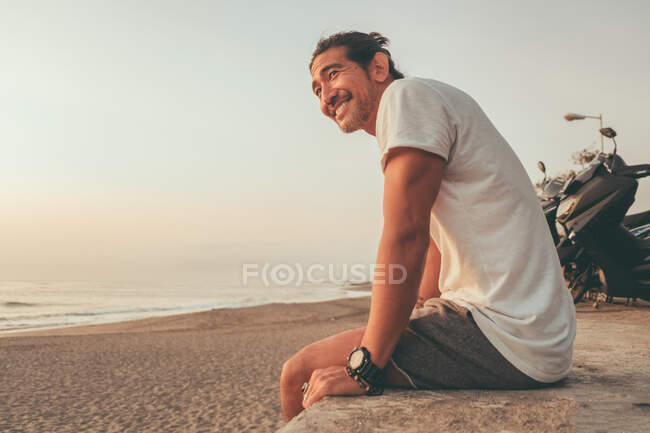 From below side view of positive active ethnic guy with motorbike sitting on stone fence and enjoying sunset on empty sandy beach — Stock Photo