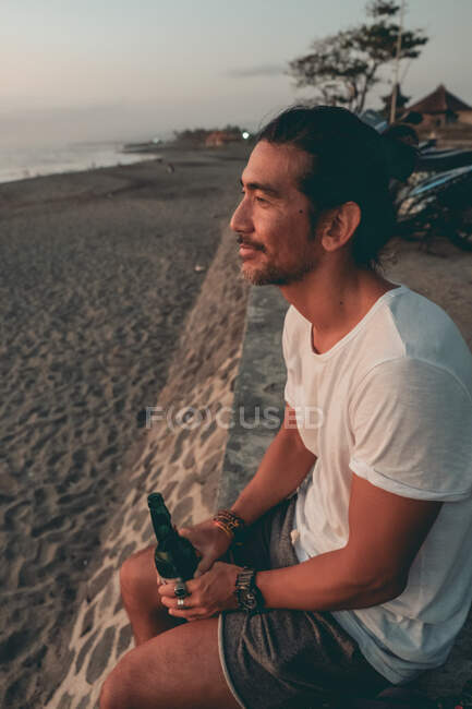 Боковой вид спокойного расслабленного молодого этнического мужчины в повседневной одежде, сидящего с бутылкой пива на песчаном пляже и наслаждающегося закатом — стоковое фото