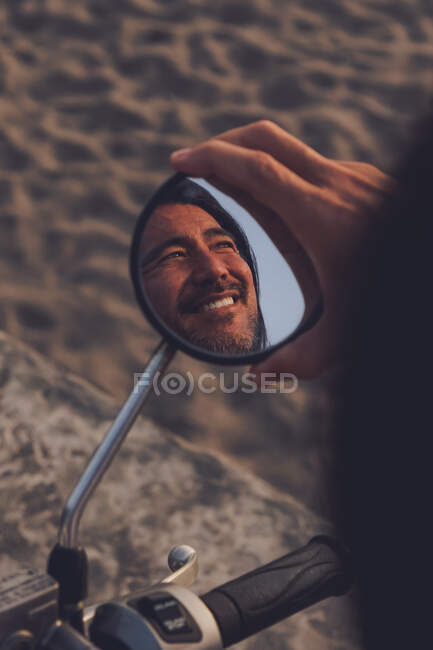 From above back view of crop joyful adult ethnic male reflected in bike mirror enjoying summertime and freedom while riding bike on sandy beach — Stock Photo