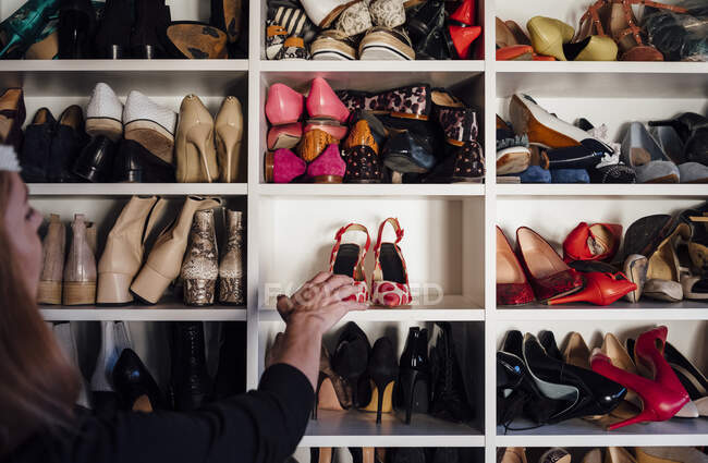 Обрізані невпізнавані жіночі руки беруть бежеві туфлі на високих підборах з полиці сучасної білої шафи — стокове фото