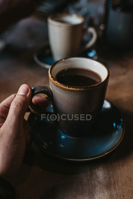 Manos de la cosecha de hembra irreconocible sentado en la mesa de madera sosteniendo café caliente en una taza rústica - foto de stock