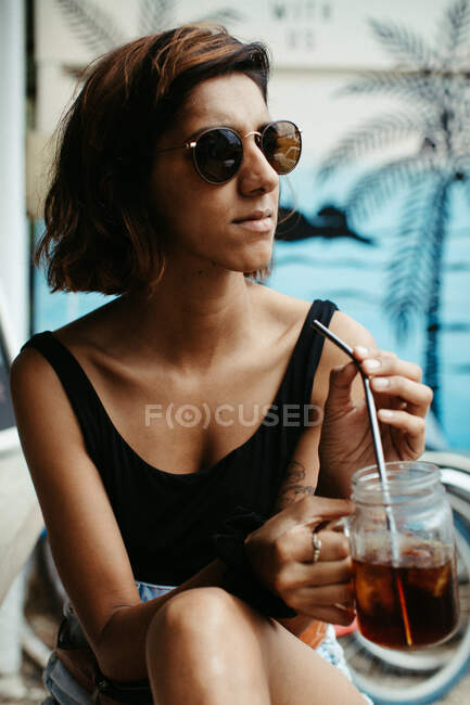 Mujer de vacaciones con gafas de sol elegantes mirando hacia otro lado sosteniendo un vaso de cóctel con vista tropical sobre fondo borroso - foto de stock