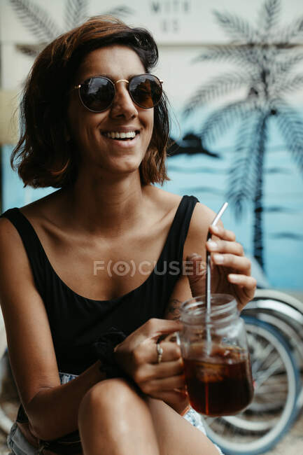 Mujer satisfecha de vacaciones con gafas de sol elegantes mirando hacia otro lado sosteniendo un vaso de cóctel con vista tropical sobre fondo borroso - foto de stock