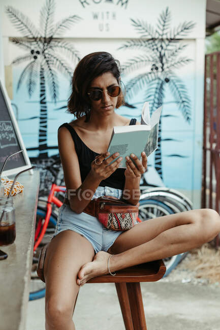 Resting woman in casual wear and trendy sunglasses reading book during refreshment in outdoors bar — Stock Photo