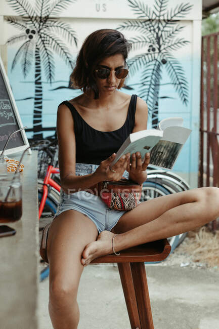 Mulher descansando no desgaste casual e óculos de sol na moda ler livro durante o refresco no bar ao ar livre — Fotografia de Stock