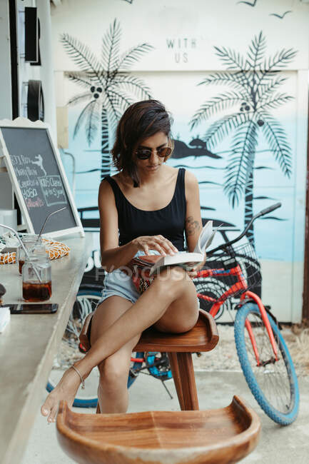 Resting woman in casual wear and trendy sunglasses reading book during refreshment in outdoors bar — Stock Photo
