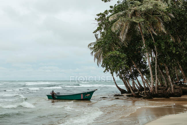 Одинокий песчаный берег с деревянной лодкой на волнистом море рядом с зелеными деревьями на фоне облачного штормового неба — стоковое фото