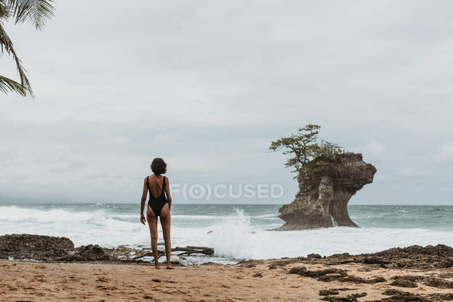 Vista trasera de la mujer en traje de baño negro caminando en la costa arenosa vacía con el océano tormentoso en el fondo nublado - foto de stock