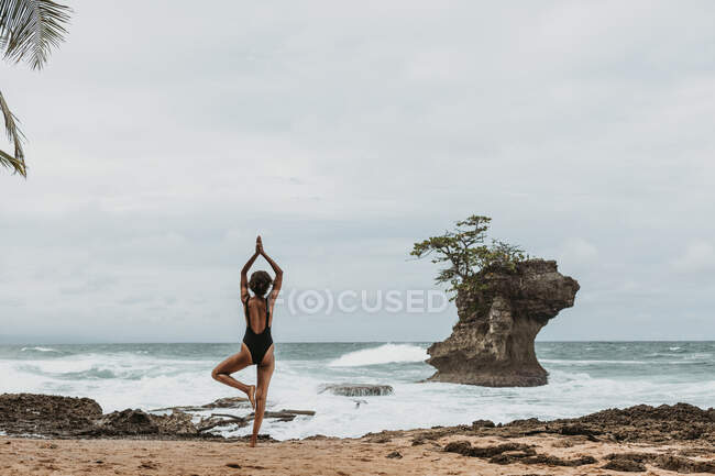 Вид ззаду безликого туриста-жінки в чорному купальнику, що балансує на одній нозі з руками, піднятими над головою на порожньому узбережжі в штормі — стокове фото