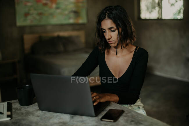 Ernsthaft fokussierte Frau tippt auf Laptop, während sie am Tisch auf einer modernen Marmorarbeitsplatte in der Tischküche sitzt — Stockfoto