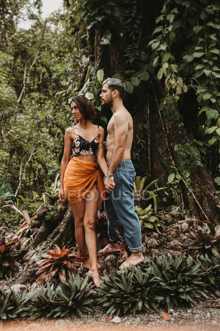 Loving sensual couple in light casual wear standing and holding hands among green tropical forest — Stock Photo