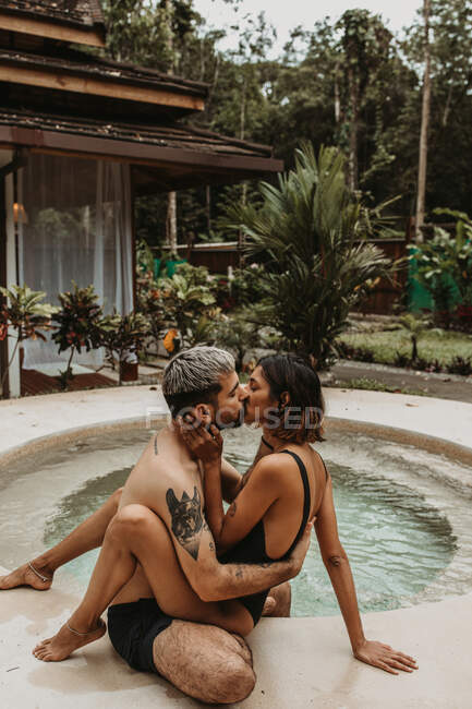 Вид сбоку на чувственную молодую пару в купальниках обнимающую и целующуюся у бассейна с тропическими зелеными растениями на заднем плане — стоковое фото