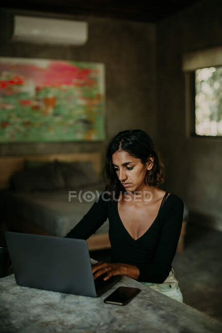 Mujer enfocada grave escribiendo en el ordenador portátil mientras está sentado en la mesa en la cocina de mesa encimera de mármol moderno - foto de stock