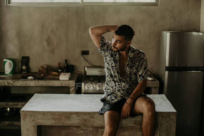 Joven pensativo con ropa casual mirando hacia otro lado y sentado en el mostrador en la cocina - foto de stock