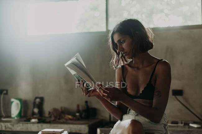 Interessato giovane donna in reggiseno e breve godendo la lettura di un libro mentre seduto con gambe incrociate sul bancone in cucina — Foto stock