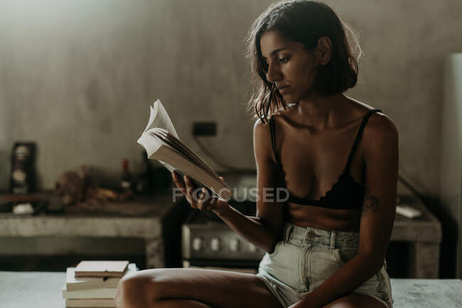 Interested youthful woman in bra and short enjoying reading a book while sitting on marble countertop in kitchen — Stock Photo