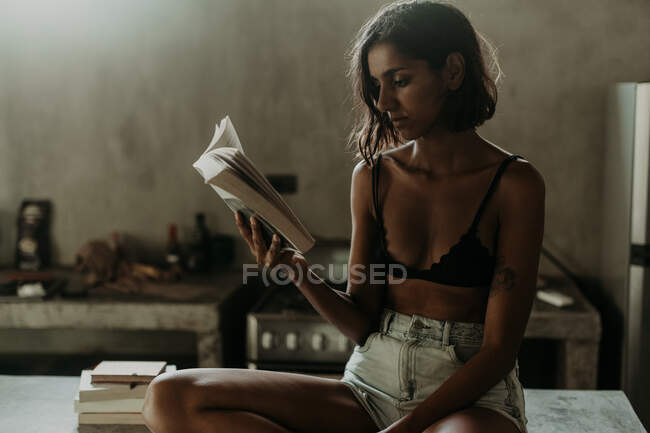 Mulher jovem interessado em sutiã e curta apreciando a leitura de um livro enquanto sentado na bancada de mármore na cozinha — Fotografia de Stock