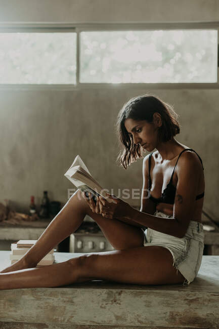 Заинтересованная молодая женщина в бюстгальтере и с небольшим удовольствием читает книгу, сидя на мраморной столешнице на кухне — стоковое фото