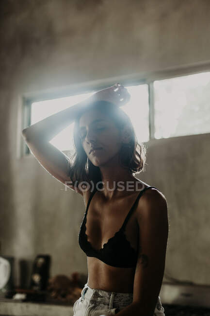 Mulher jovem macia em sutiã e com os olhos fechados levantando a mão para cima e sentado na cozinha — Fotografia de Stock