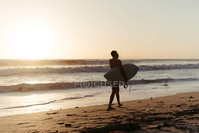 Vista posterior de la silueta del hombre sosteniendo la tabla de surf mientras camina a lo largo de la orilla del mar de arena en verano durante el atardecer - foto de stock