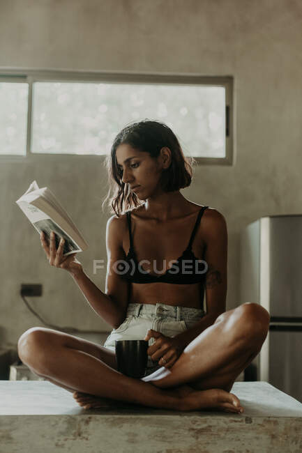 Interessante junge Frau im BH und kurzer Freude am Lesen eines Buches, während sie in der Küche auf einer Marmorarbeitsplatte sitzt — Stockfoto