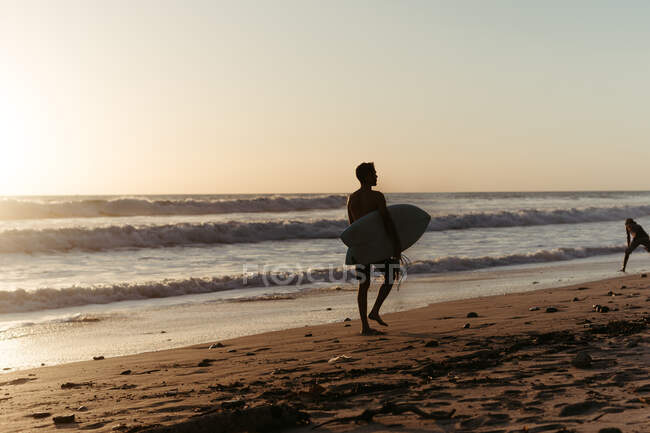 Вид ззаду на силует людини, що тримає дошку для серфінгу під час прогулянки вздовж піщаного узбережжя влітку під час заходу сонця — стокове фото