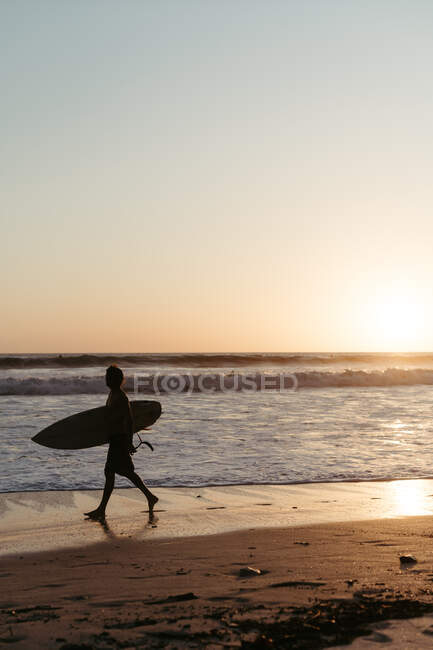 Вид збоку силует людини тримає дошку для серфінгу під час прогулянки вздовж піщаного узбережжя в літній час під час заходу сонця — стокове фото