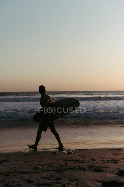 Vista lateral de la silueta del hombre sosteniendo la tabla de surf mientras camina a lo largo de la orilla del mar de arena en verano durante el atardecer - foto de stock