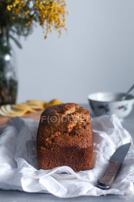 Delicioso y apetitoso pastel casero de limón y semillas de amapola colocado en tela blanca con cuchillo de cocina en la mesa con un tazón y flores de mimosa en el fondo - foto de stock