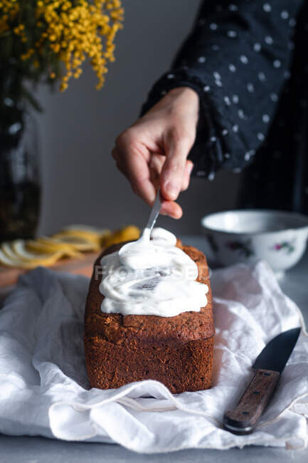 Mão de colheita de dona de casa decorando delicioso bolo de esponja de limão e papoula caseiro com chantilly à mesa com buquê de flores de mimosa no fundo — Fotografia de Stock