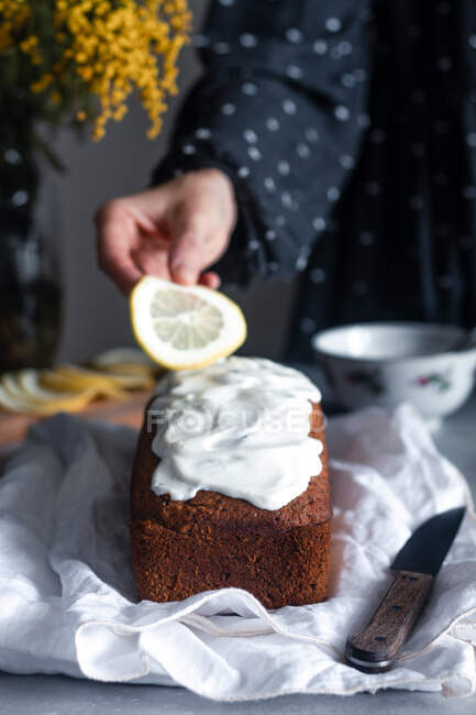 Mão de corte de mulher colocando fatia de limão no delicioso bolo caseiro fresco coberto com chantilly colocado em pano branco na mesa da cozinha com buquê de flores de mimosa no fundo — Fotografia de Stock