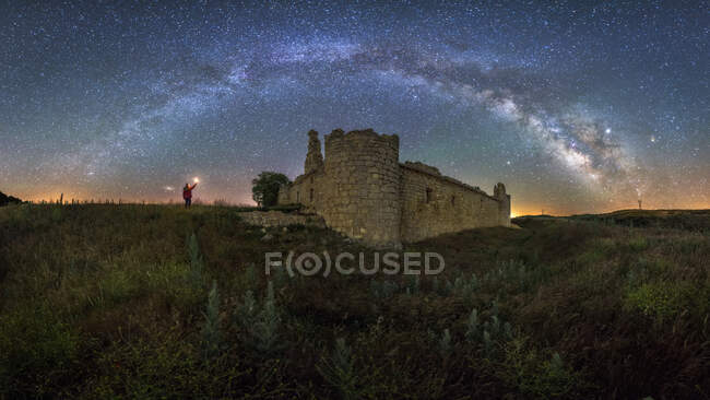 Desde abajo vista trasera de un turista anónimo con linterna explorando el viejo castillo en ruinas bajo la Vía Láctea en la noche estrellada - foto de stock