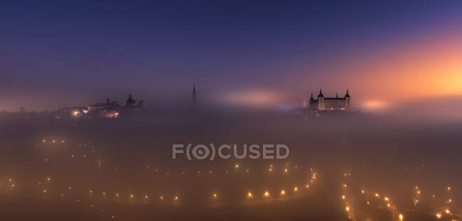 Desde arriba asombroso paisaje de ciudad medieval iluminada y palacio Alcázar de Toledo en nebuloso amanecer colorido - foto de stock