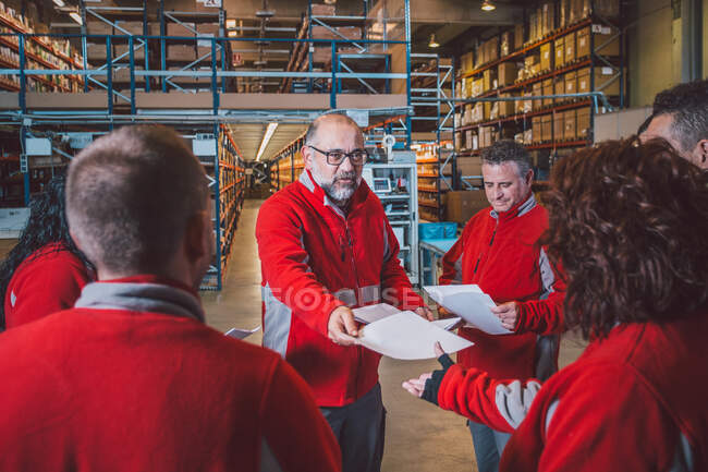 Barbuto senior manager maschile dando istruzioni sui documenti al gruppo di dipendenti in uniforme rossa i magazzino moderno — Foto stock
