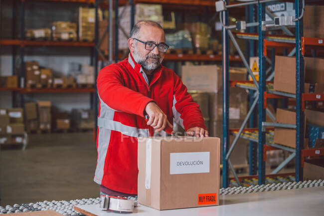 Senior bearded male worker in red uniform opening parcel with paper knife and looking at camera in big storage — Stock Photo