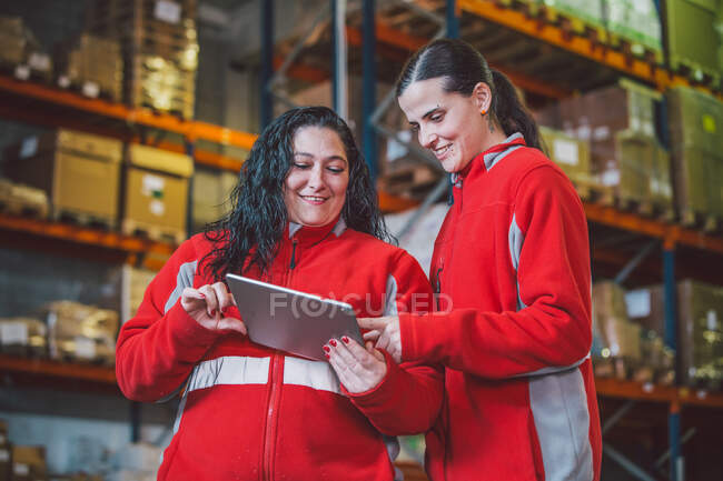 D'en bas de jeunes femmes en uniforme rouge souriant tout en utilisant une tablette numérique pendant le travail dans un entrepôt moderne — Photo de stock