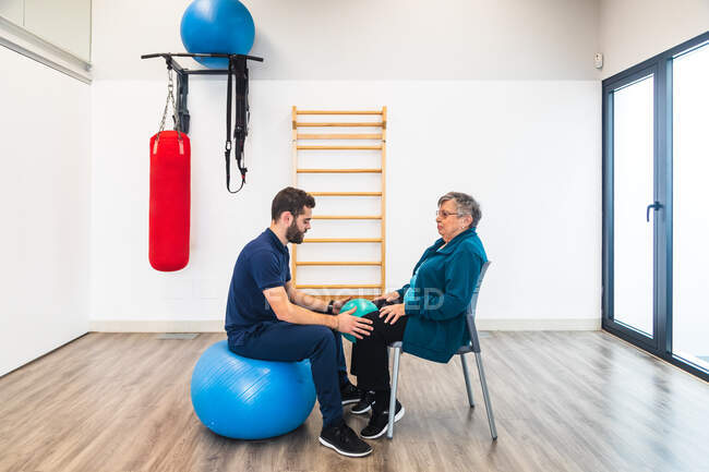 Vista lateral del entrenador masculino barbudo sentado en la pelota de ejercicio delante de la señora mayor en la silla con una pequeña bola entre las rodillas en el gimnasio - foto de stock