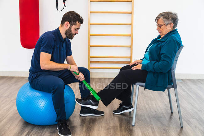 Vue latérale de l'entraîneur personnel masculin assis sur la balle bleue d'exercice tout en utilisant la bande élastique verte sur la cheville de la vieille femme dans la salle de gym — Photo de stock