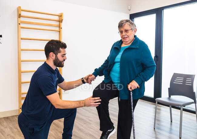 Instructeur masculin soutenant la vieille femme utilisant le train dans la salle de gym — Photo de stock