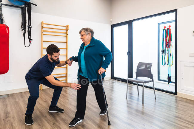 Instrutor masculino apoiando mulher idosa usando trem no ginásio — Fotografia de Stock
