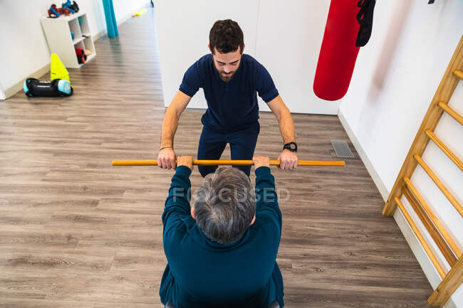 Back view from above of senior lady holding stick with male personal trainer during physiotherapy workout in gym — Stock Photo