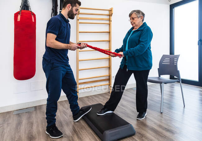 Seitenansicht einer alten Frau und eines männlichen Fitnesstrainers, die sich gegenüberstehen und mit einer Trittplattform in der Turnhalle Gummibänder ziehen — Stockfoto
