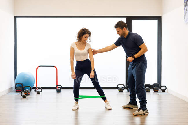 Junge Frau in aktiver Kleidung steht mit grünem Gummiband an den Beinen und trainiert unter Aufsicht eines männlichen Trainers im Fitnessstudio — Stockfoto