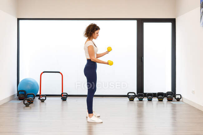 Vue latérale de la femme concentrée en tenue de sport debout et faisant des boucles biceps avec des haltères jaune clair dans la salle de gym moderne — Photo de stock