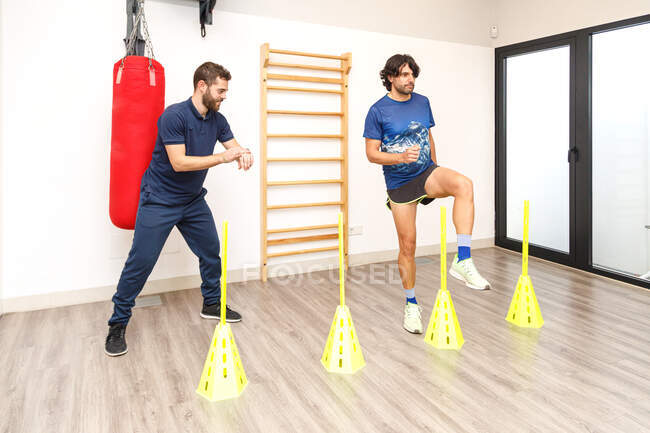 Junger Mann in aktiver Kleidung steht auf einem Bein, während er mit gelben Kegeln auf Geschwindigkeit trainiert, wobei der Trainer lächelt und die Zeit auf der Armbanduhr im Fitnessstudio überprüft — Stockfoto