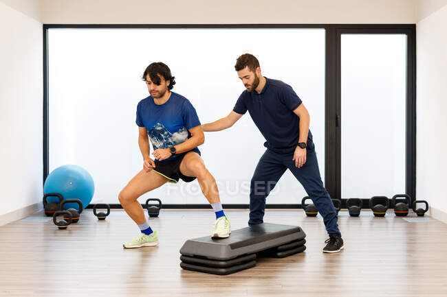 Sportler in aktiver Kleidung führt Kniebeugen mit einem Bein auf Trittplattform durch, während männliche Trainer den Prozess im Fitnessstudio steuern — Stockfoto