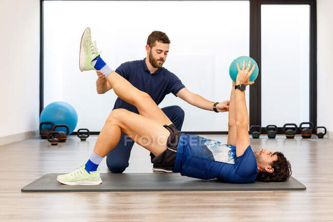 Side view of man in active wear lying on mat with one raised leg and hands with small ball under control of fitness trainer in gym — Stock Photo