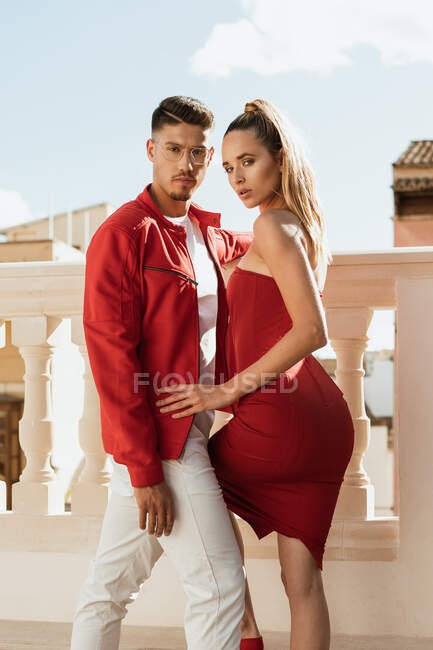 Seitenansicht des emotionslosen männlichen Modells in stylischer roter Jacke und des weiblichen Modells im trendigen roten Kleid, das auf dem Balkon steht und in die Kamera blickt — Stockfoto