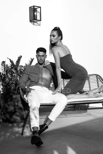 Модная пара модель в модной одежде, сидящая на диване и смотрящая в камеру на открытой террасе при солнечном свете — стоковое фото