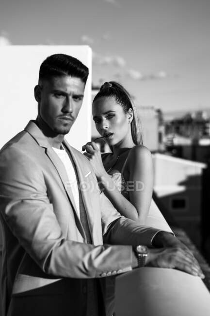 Black and white side view of unemotional couple in trendy suit and dress standing on balcony and looking at camera — Stock Photo